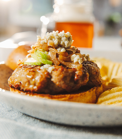 A open-faced pork burger sits on a grey plate with a side of ruffle potato chips. The burger is topped with slices of green apple, caramelized onions and crumbled blue cheese.