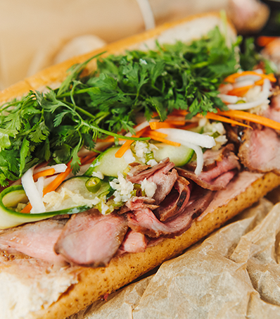 A fully loaded Vietnamese Banh Mi sandwich sits diagonally on brown paper. The sandwich is on a baguette roll and there is plenty of slices of beef, lots of pickled cucumbers, carrots and daikon, and lots of fresh cilantro leaves.