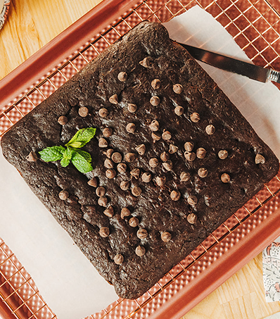 A square brownie rests on top of a piece of wax paper and a rose gold cooling rack. The brownie is garnished with chocolate chips and a single sprig of mint.