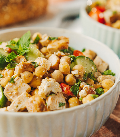 A small white bowl is filled with a salad consisting of cooked, cubed chicken, diced cucumbers, cherry tomatoes, chickpeas and seasonings. The salad is garnished with a sprig of mint and served with a loaf of fresh bread.