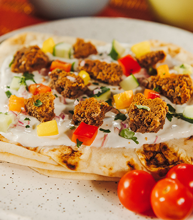 A pite rests on a light coloured plate. the pita is topped with white sauce, torn pieces of falafel, diced cucumbers and red and yellow peppers.