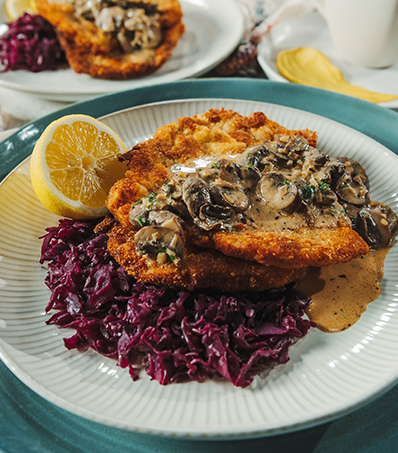 A light coloured plate rests on top of a blue serving platter. The light coloured plate has a crispy pork chop resting atop of shredded purple cabbage and is dressed in a mushroom gravy sauce. The plate has a half lemon garnish and a simillar plate can be seen in the background behind the first.
