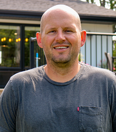 A portrait-style photo of a bald man wearing a grey t-shirt. He is standing in the sunshine in front of his back deck with his house slightly visible in the background.