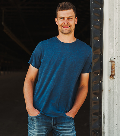 Portrait of a man wearing a deep blue t-shirt and jeans. The man is standing in front of a open but obscured door to an empty farm building with his hands in his pocket.