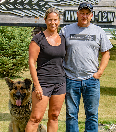 Portrait of a woman and a man standing outside on a sunny day. Part of a wooden sign can be seen behind them and their Shepard dog sits beside them.