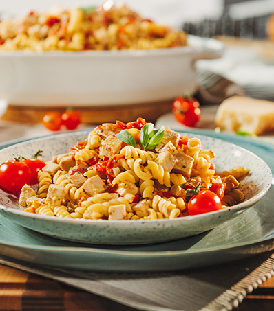 A single serving of turkey, tomato and boursin pasta garnished with cherry tomatoes sits in a shallow bowl. An out of focus family-sized portion of the same pasta dish can be seen in the background.