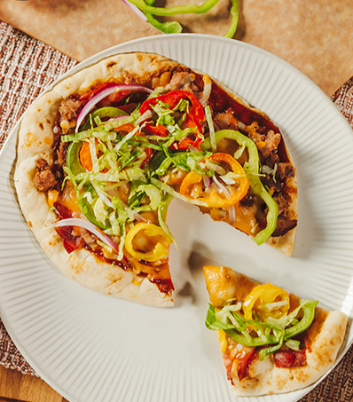 A flat bread pizza sits on a circular white plate. The flatbread is topped with BBQ sauce, chopped cooked turkey burgers, turkey pepperoni, marble cheese and is garnished with shredded lettuce and pickled pepper rings.