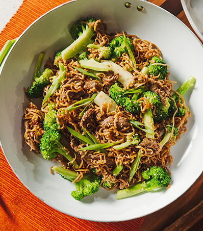 A top down perspective photo of a stir fry of ramen noodles, slices of beef and broccoli florets in a white pan. The stir fry is garnished with slices of green onion.