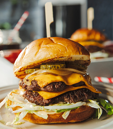 A tall double patty smash burger with cheese and a golden toasted bun sits on a light coloured plate. The burger is topped with shredded iceberg lettuce and pickle coins and is held together with a wooden skewer. Another smash burger is out of focus in the background.