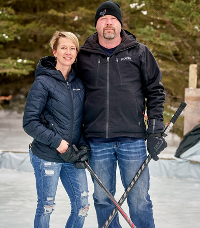 A man and a women stand beside one another on an ice rink with out of focus trees in the background. They are both wearing dark puffer jackets and holding hokey sticks.
